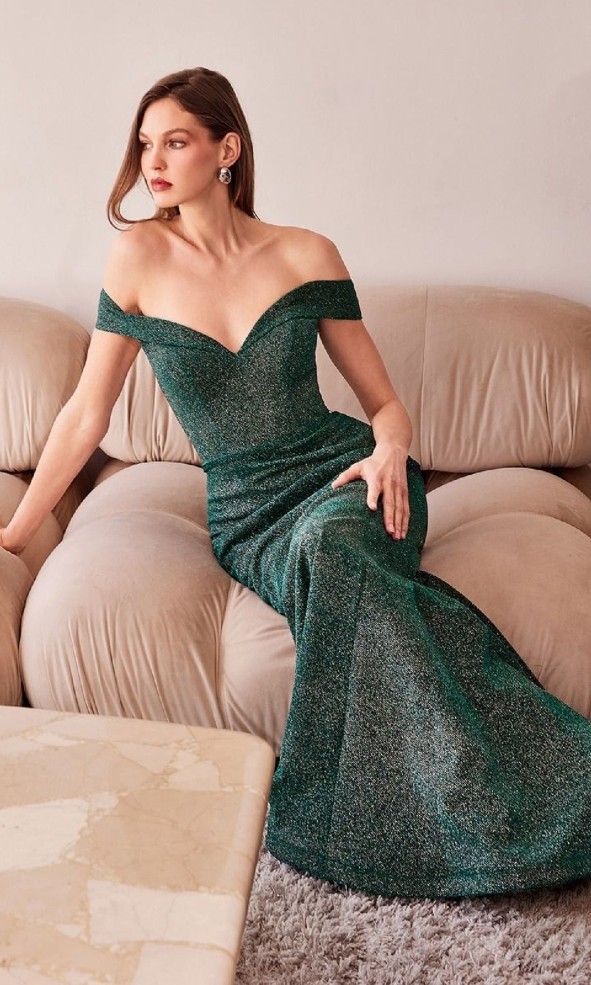 A woman in the Off-Shoulder Sweetheart Prom Gown: Amoris T602 sits on a beige sofa, looking to the side with one arm resting on the couch.